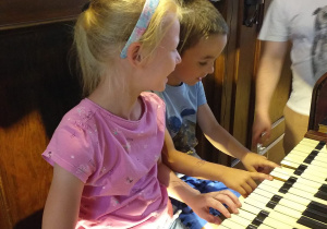 dwie dziewczynki siedzą przy organach, naciskają klawisze, obok nich stoi pan od rytmiki